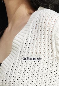 Bílý pletený cardigan s výstřihem do V, zdobený texturovaným vzorem a tmavě modrým logem "adidas" na levé straně.
