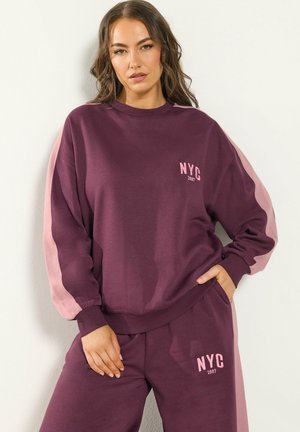 Frau trägt einen lila Trainingsanzug mit pinken Streifen, Hände in den Taschen, "NYC 1987" auf Sweatshirt und Hose gedruckt, steht vor einer hellen Wand.