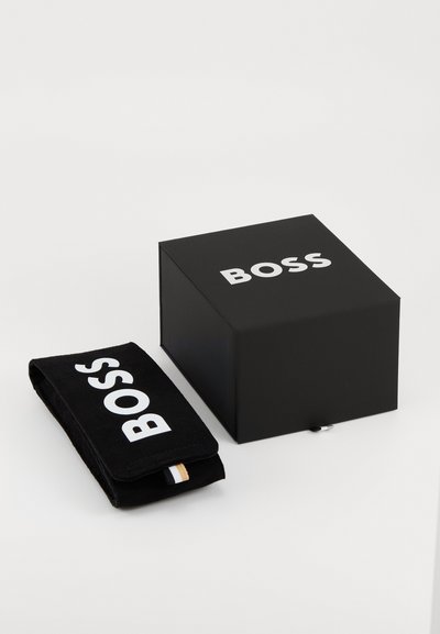 Boîte noire avec le logo "BOSS" blanc ; à côté, un sac en velours noir avec le logo et une petite bordure rayée en bas. Texture lisse.