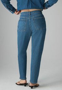 Blå denimjeans med en avsmalnande passform, två bakfickor och fransar vid fållarna. Materialet har en texturerad yta och medeltonad tvätt.
