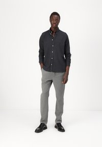 Donkergrijs overhemd met lange mouwen, gecombineerd met lichtgrijze broek en zwarte schoenen, staand tegen een effen witte achtergrond.