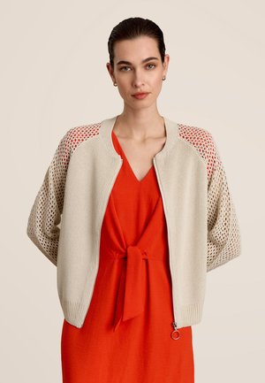 Beige gebreide cardigan met opengewerkte patroon mouwen en ritssluiting, gedragen over een rode jurk met een gestrikte knoop in de taille.