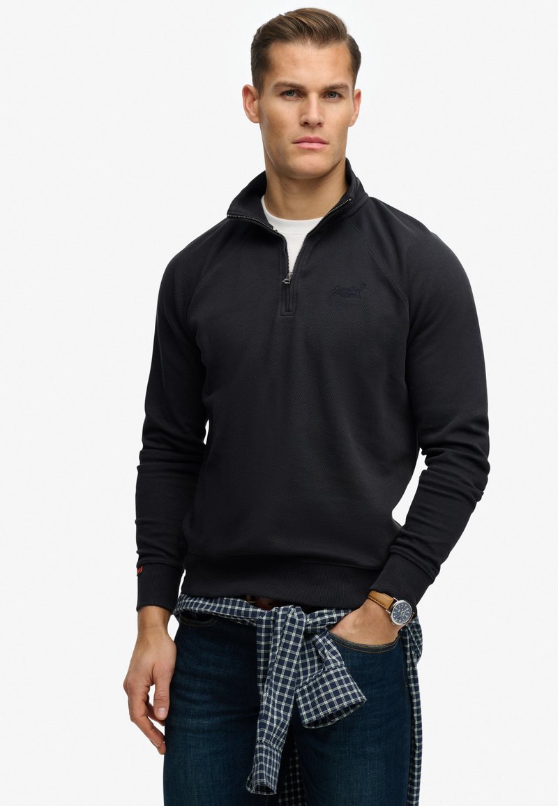 Homme portant un pull noir à demi-fermeture éclair, un sous-maillot blanc, un jean foncé, une chemise à carreaux nouée à la taille et une montre-bracelet, debout devant un fond blanc.