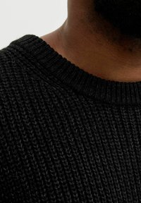 Nahaufnahme des Nackens und der Schulter einer Person, die einen schwarzen gerippten Strickpullover mit einem Rundhalsausschnitt trägt.