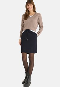 Hellbrauner V-Ausschnitt-Pullover, schwarzer Sportrock mit Kordelzug, schwarze Strumpfhose und schwarze Schnürstiefel. Einfaches, lässiges Outfit.