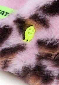 Tissu en fausse fourrure rose avec des taches foncées, présentant un petit accessoire en plastique vert en forme de visage souriant qui fait un clin d'œil.