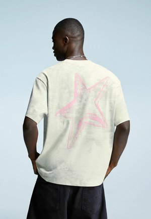 Man wearing off-white T-shirt with large pink star design on the back and black shorts, standing against a plain light blue background.