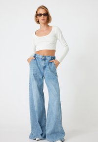 Weißes, geripptes, langärmeliges Cropped-Top kombiniert mit weit geschnittenen, hellblauen Denim-Jeans mit hoher Taille und großen Seitentaschen.