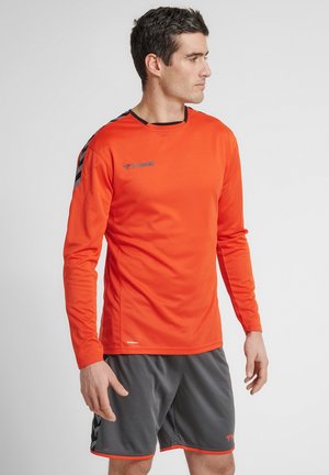 Mand iført en skarp orange langærmet sportstrøje og mørkegrå træningsshorts, stående og kiggende mod højre mod en ensfarvet baggrund.