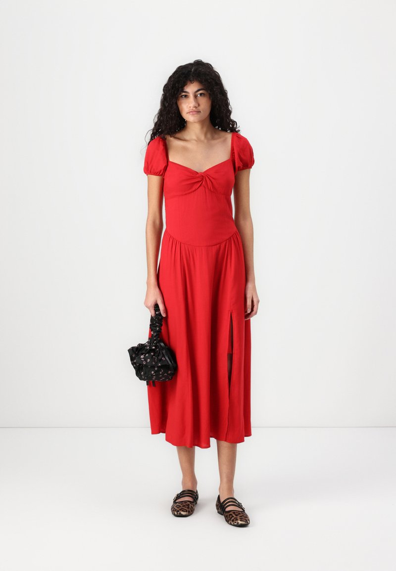 Rotes, knielanges Kleid mit kurzen Puffärmeln, einer gedrehten Detail am Ausschnitt und einem seitlichen Schlitz. Das Model hält eine schwarze Handtasche mit strukturiertem Design.