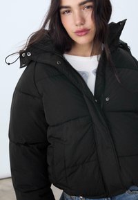 Giacca a vento nera con colletto alto, chiusura con zip e bottoni a pressione, e tasche laterali. Il tessuto sembra liscio con un design trapuntato.