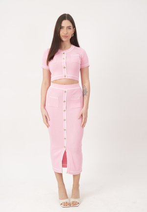 Kobieta stojąca, ubrana w różowy komplet dwuczęściowy z złotymi guzikami, krótkimi rękawami, krótkim topem i spódnicą midi z przednim rozcięciem i kieszeniami.