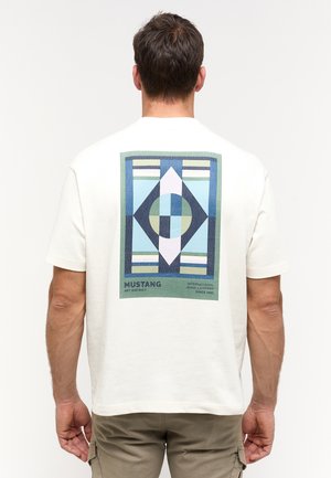 Homme portant un t-shirt blanc avec un motif géométrique bleu et vert et le texte "Mustang" dans le dos, dos à la caméra.
