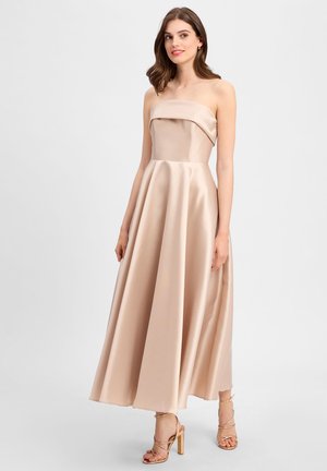 Strapless beige jurk van satijn, met een aansluitend lijfje en een uitlopende, vloerlange rok. Metallic strappy hakken complementeren de look.