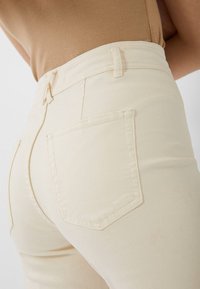 Beige korkeavyötäröiset farkut, joissa on sileä pinta, takataskut ja vyölenkit. Ominaisuuksiin kuuluu vartalonmyötäinen siluetti ja hienovaraiset ompeluyksityiskohdat.