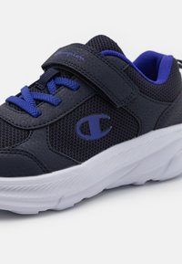Baskets de sport navy avec des accents violets, des panneaux en tissu en maille, une bande Velcro et une semelle extérieure en caoutchouc blanc, arborant un logo sur le côté.