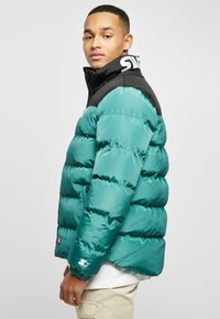 Teal puffer jacket with a black shoulder area, zip front, and ribbed cuffs. Features logo on collar and white star accent on sleeve.