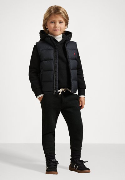 Colete acolchoado preto sobre um hoodie preto, suéter de gola alta branca, calças de treino pretas e sapatilhas pretas com detalhes em castanho. Logótipo vermelho notável no colete.