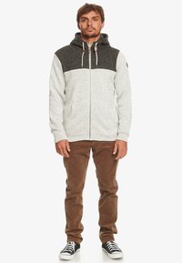 Sudadera con capucha y cremallera en gris claro con panel superior en gris oscuro. Presenta cordones y un parche de logo, combinada con pantalones de pana marrones.