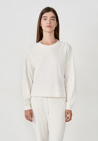 Felpa di colore crema con vestibilità comoda, scollo a girocollo e maniche lunghe. Texture liscia con polsini e orlo a coste. Abbinata a pantaloni coordinati.