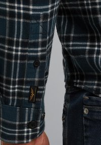 Geruite langemouwen shirt manchet met zwarte knopen en een kleine zwart-gouden patch naast de zakdetails van donkerblauwe denim jeans.