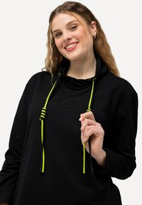 Sudadera con capucha negra que presenta un logo texturizado, cordones amarillos brillantes y un corte relajado. El material parece suave y cómodo.