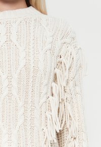Cremfarbener Strickpullover mit Kabelmustern, ausgestattet mit langen Fransen entlang der Ärmel. Strukturiertes Material mit einem gerippten Kragen.
