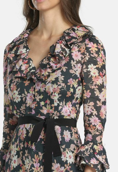 Femme portant une robe noire à fleurs roses et blanches, avec un col et des poignets volantés, ainsi qu'une ceinture noire nouée à la taille.