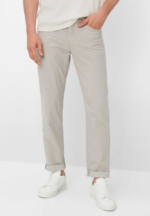 Uomo che indossa pantaloni grigio chiaro aderenti con orli arrotolati, sneakers bianche e una camicia bianca strutturata infilata, in piedi con una mano in tasca.