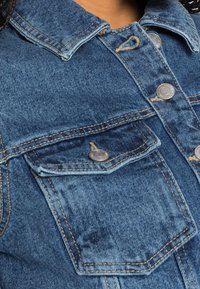 Kurtka dżinsowa w jasnoniebieskim kolorze, z metalowymi srebrnymi guzikami i dwoma kieszeniami na piersi, podkreślająca teksturowaną tkaninę oraz wzmocnione szwy.