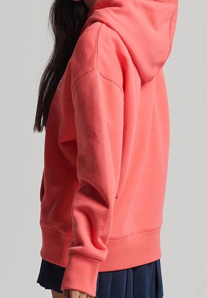 Huppari korallinvärisenä, valmistettu pehmeästä kankaasta. Rentouttava istuvuus, pitkät hihat ja kengurulokero. Näkyvä resorireuna ja -hihansuut.