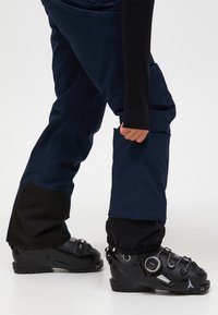 Marineblå skibukser med sorte nederste accenter, har lommer og en løs pasform, parret med sorte skistøvler med justerbare stropper og et strømlinet design.