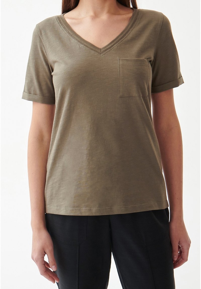 Femme portant un t-shirt à manches courtes vert olive à col en V avec une poche sur la poitrine et un pantalon noir, debout devant un fond blanc.
