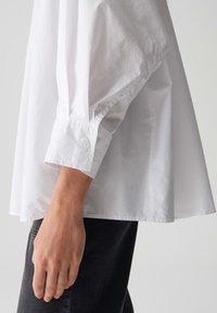 Witte blouse met lange mouwen en een manchet met knoop, zichtbaar op een arm die donkere broek draagt, tegen een effen lichte achtergrond.