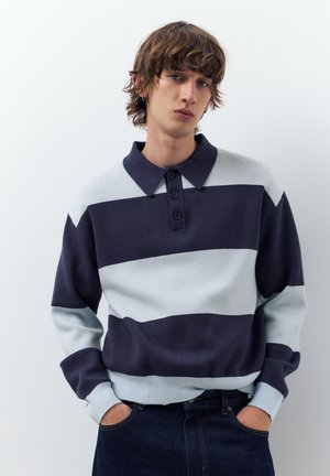 Mladi moški z valovitimi rjavimi lasmi, oblečen v mornarsko modro in svetlo sivo polo majico z dolgimi rokavi s črtami ter temne kavbojke, z rokami v žepih, nevtralen izraz na obrazu.