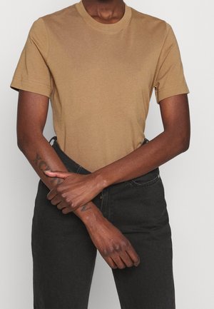 Persoon draagt een beige T-shirt met korte mouwen en zwarte broek, met de armen over elkaar ter hoogte van de taille, waarbij tatoeages op de linker onderarm zichtbaar zijn.