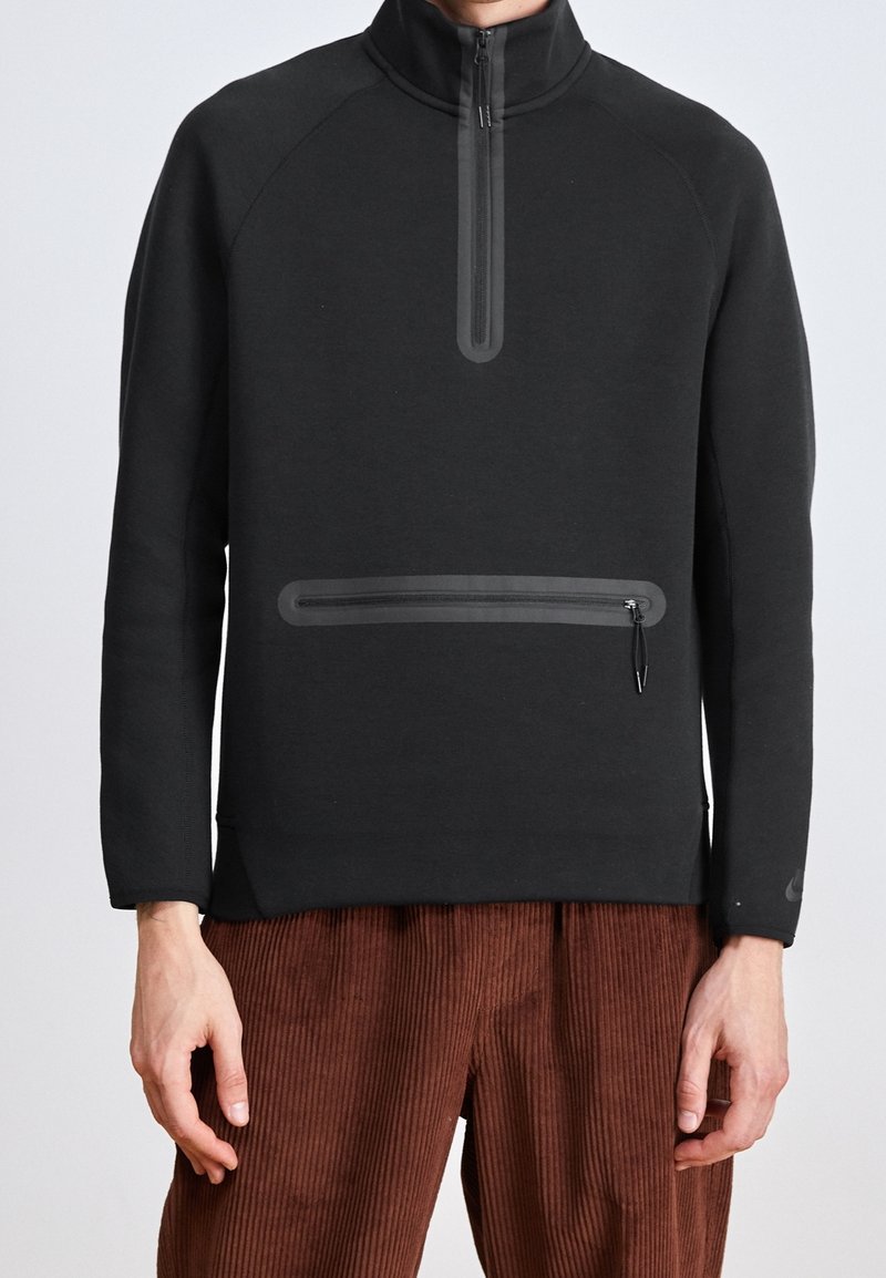 Sudadera negra de cuello alto y cremallera de cuarto. Presenta un bolsillo con cremallera y tejido texturizado. Combinada con pantalones de pana marrones.