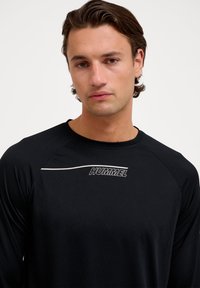Juodas ilgomis rankovėmis sportinis marškinėliai, pagaminti iš tekstūruoto audinio. Pasižymi baltu "HUMMEL" logotipu ir kontrastinga linija ant krūtinės.