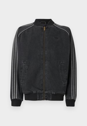 Uomo con capelli corti e ricci che indossa una giacca di jeans nera con scritta "adidas" in rilievo sulla schiena, in piedi davanti a uno sfondo semplice.