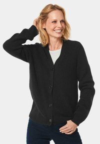Schwarze Strickjacke mit V-Ausschnitt, mit einem strukturierten Muster, gerippten Bündchen und fünf Knöpfen vorne. Über einem weißen Oberteil getragen.