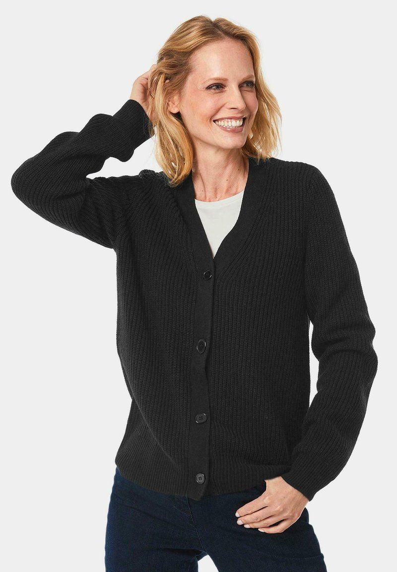 Schwarze Strickjacke mit V-Ausschnitt, mit einem strukturierten Muster, gerippten Bündchen und fünf Knöpfen vorne. Über einem weißen Oberteil getragen.