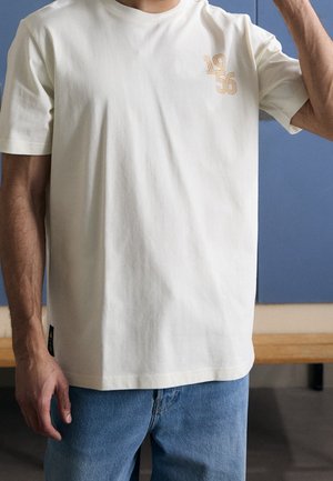 Mann trägt ein schlichtes, cremeweißes T-Shirt mit kurzen Ärmeln und der Aufschrift "1956" auf der Brust sowie eine blaue Jeans. Er steht drinnen vor einer blauen Wandverkleidung.