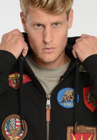 Zwarte zip-up hoodie met geborduurde patches in rood, blauw en beige. Heeft een trekkoord capuchon en een metalen rits met leren trekker.