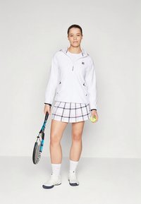 Hvit jakke med glidelås og svarte og rosa detaljer, rutete skjørt i hvit og lys rosa, holder en tenniskølle og ball, iført hvite sneakers.