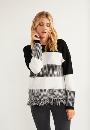 Donna con lunghi capelli biondi che indossa un maglione a righe chevron bianco e nero con frange sull'orlo, che posa con gli occhi chiusi e la mano vicino al viso.