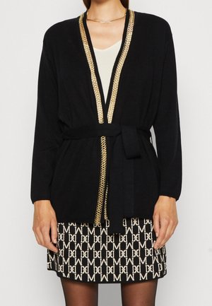 Schwarzer Cardigan mit einem goldenen Kettendetail entlang der offenen Vorderseite, mit langen Ärmeln und einem Bindegürtel. Getragen über einem gemusterten schwarzen und weißen Rock.