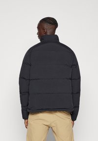Veste noire matelassée avec un col haut, présentant une forme carrée et des coutures horizontales. Le matériau semble lisse et durable.