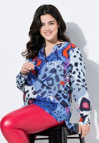 Farverig langærmet top med et dristigt abstrakt print i blå, rød og sort. Modellen bærer skinnende røde leggings og holder briller.