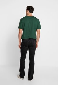 Grünes Baumwoll-T-Shirt mit Rundhalsausschnitt, kombiniert mit schwarzen Denim-Jeans. Die Jeans haben eine standardmäßige Passform mit Gesäßtaschen und minimaler Ausstattung.