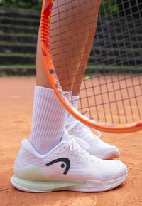 Hvite tennissko med teksturerte overflater, grønne detaljer og svart logo. Brukt med hvite ribbestrikkede sokker på en leirbane. Tennist racket synlig.
